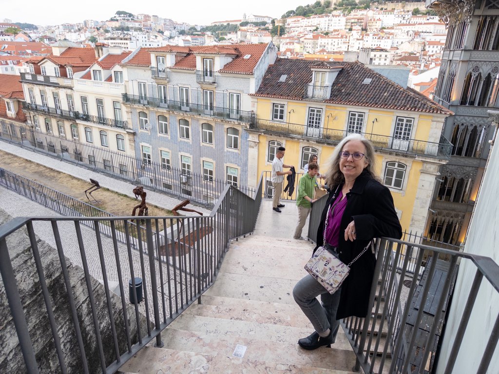 View down outdoor staircase of a smiling woman with white and gold-streaked hair, wearing glasses, black sweater and a white handbag leaning against a railing, with colorful Lisbon buildings in the background.